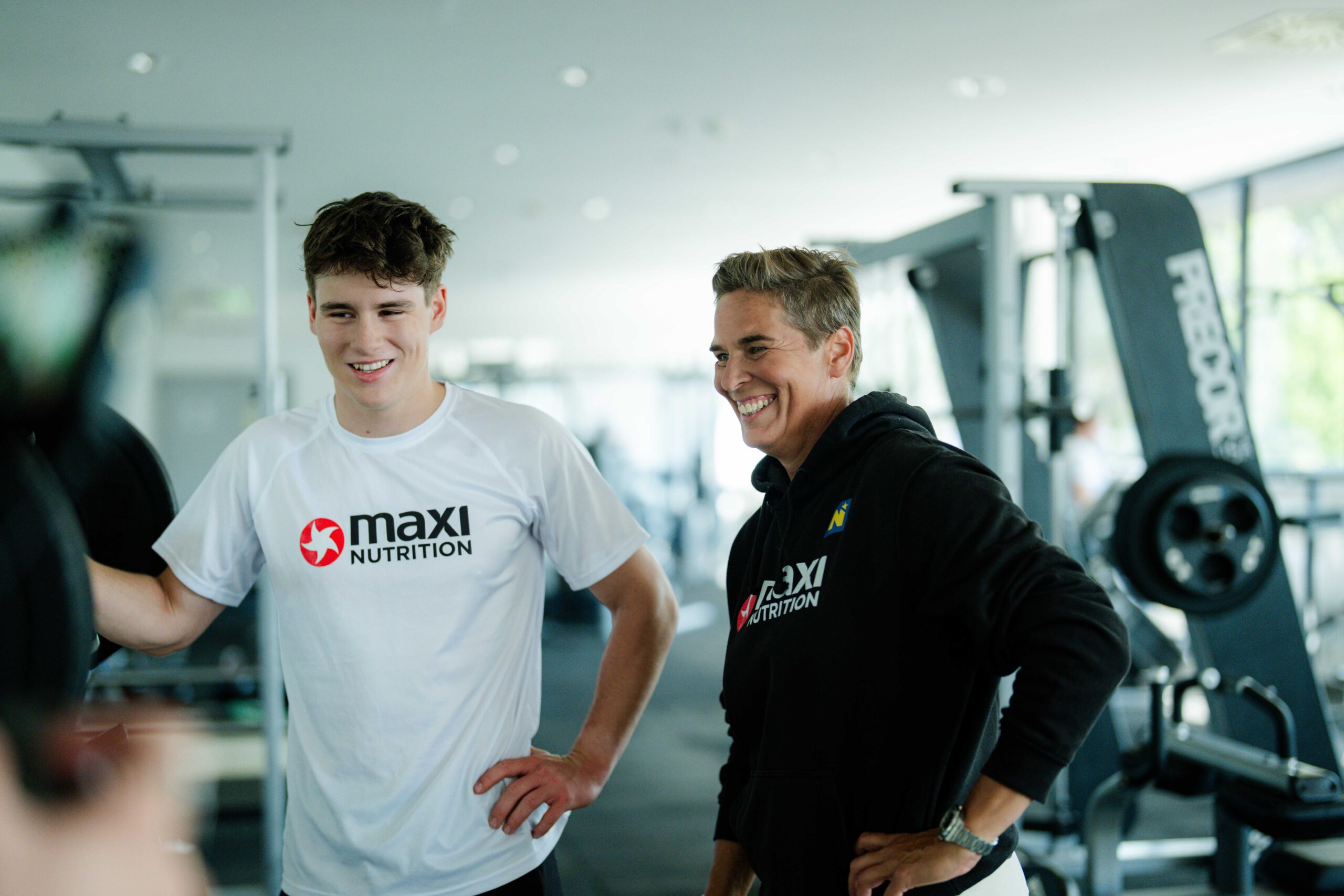 Elias und Angelika stehen gemeinsam im Fitnessstudio. Sie blicken beide nach unten links und lachen. Man sieht, dass sich die beiden gerade unterhalten und über etwas lachen. Beide haben ihren Arm in die Hüfte gestützt.
