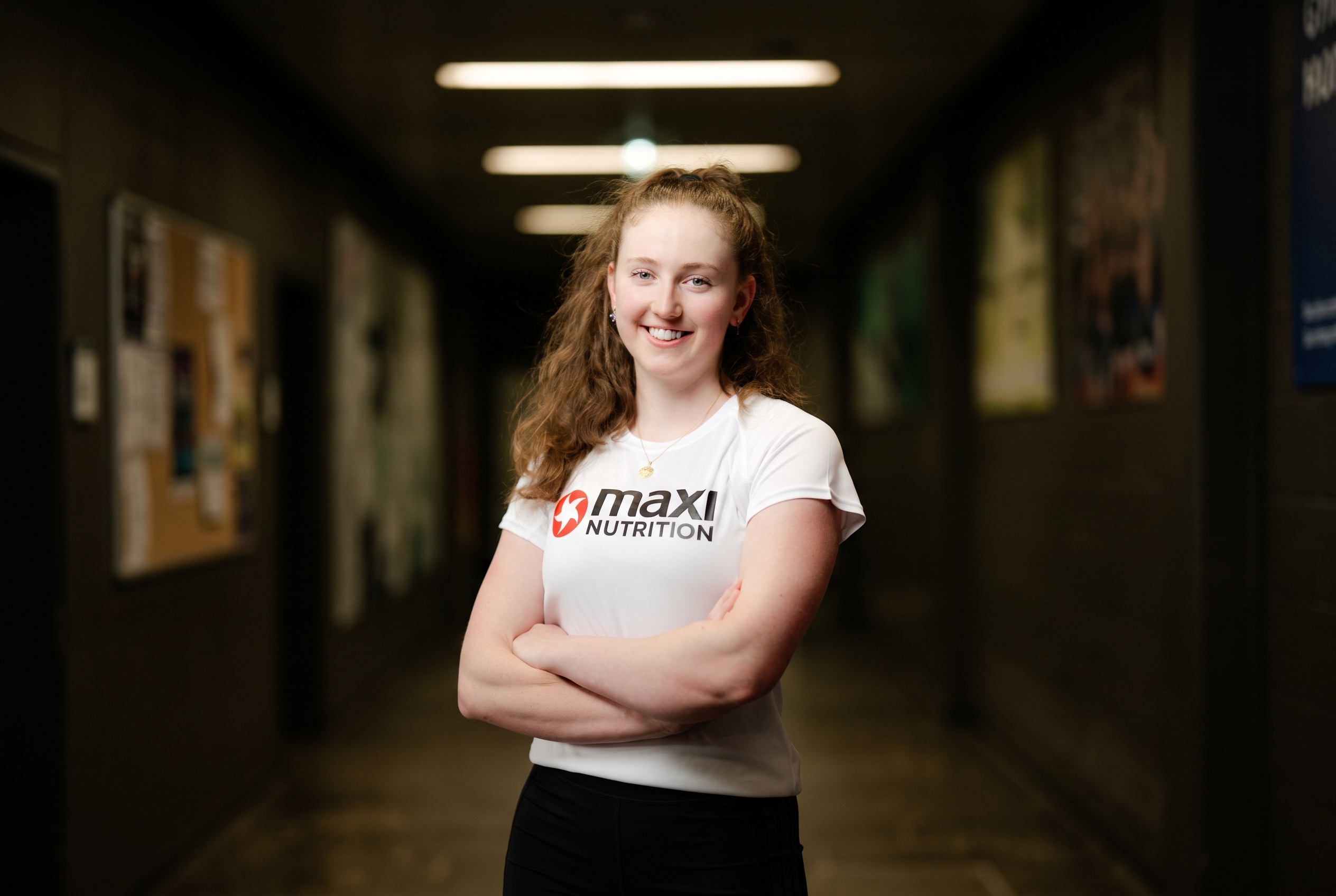 Angelika trägt ein Kurzarm-Shirt und hat die Arme vor der Brust verschränkt. Sie steht etwas schräg und lächtelt mit Zähnen in die Kamera. Sie trägt einen Pferdeschwanz und hat lockige, lange Haare.