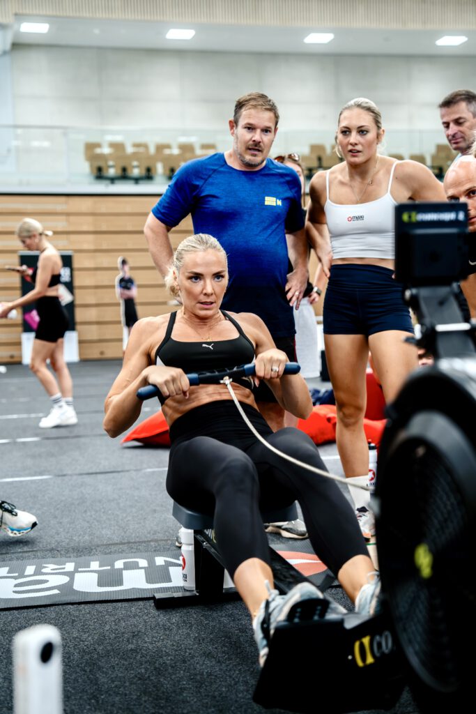 Ivona Dadic blickt konzentriert auf das Display des Rudergeräts und zieht an der Stange. Hinter ihr stehen zwei Personen und beobachten die ehrgeizige Sportlerin.