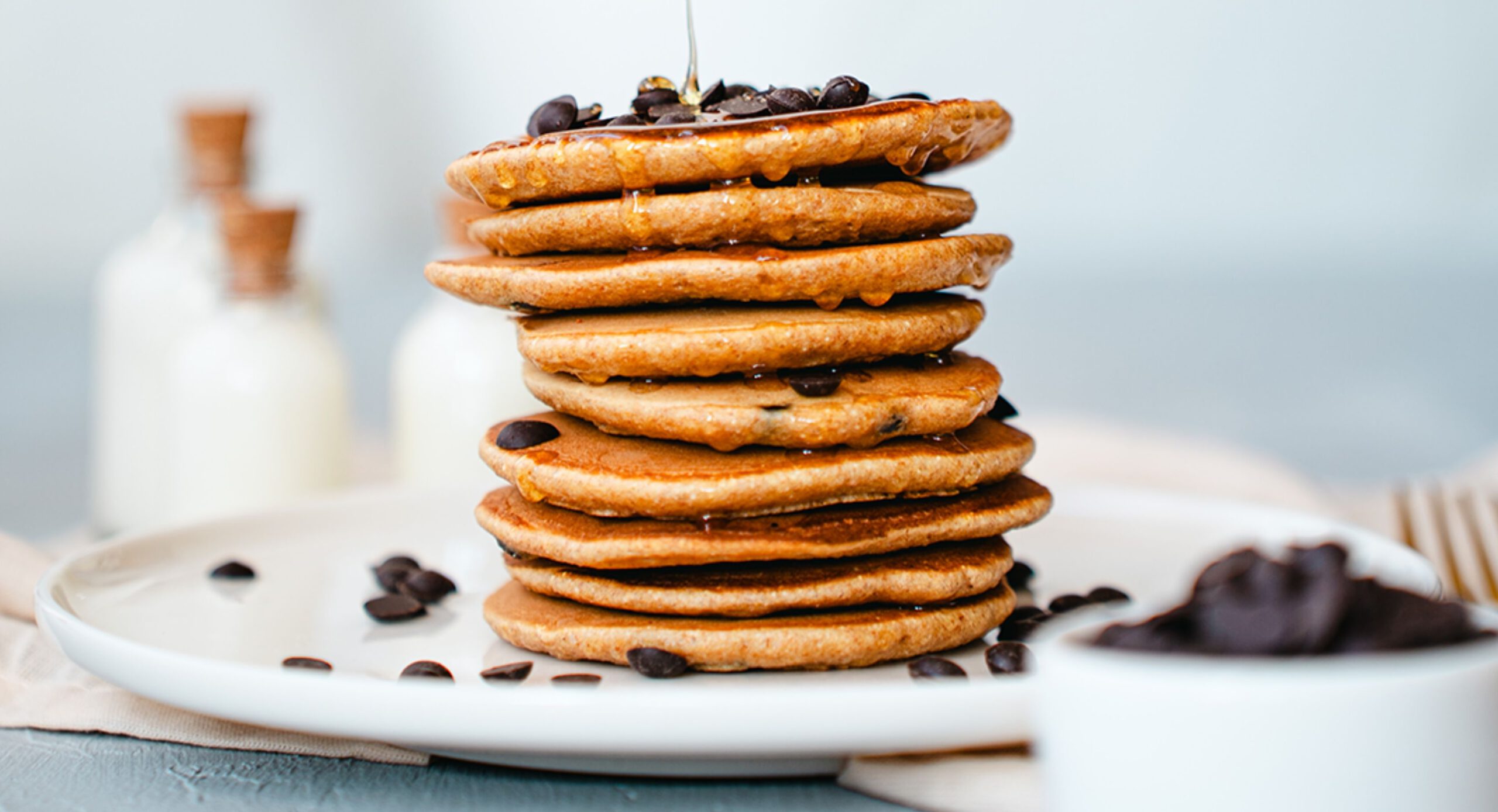 Ein Stapel Protein-Pancakes mit Choco-Chips obendrauf auf einem Teller angerichtet. Daneben steht eine kleine Schüssel gefüllt mit Choco-Chips.