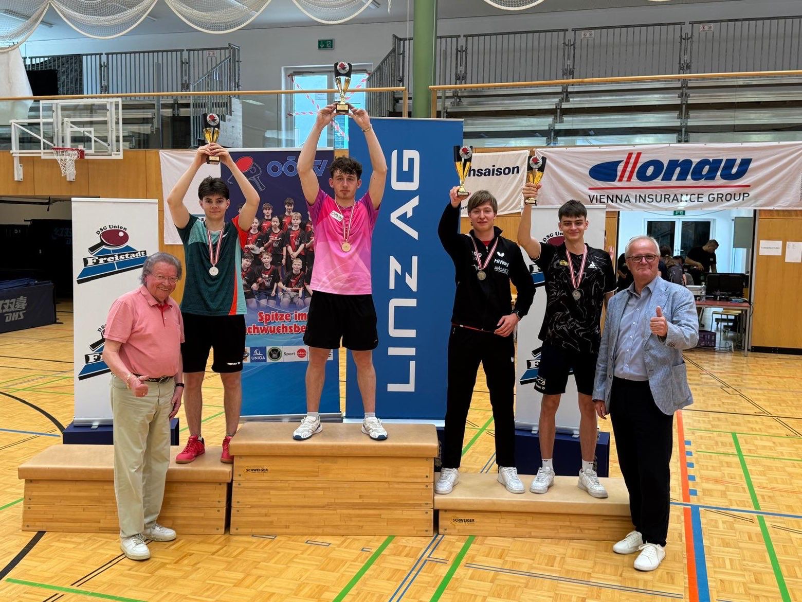 Julian Rzihauschek steht auf einem Podest und hält den Pokal in die Höhe. Neben ihm stehen ebenfalls auf einem Podest weiter unten Platz 2 und die beiden Platz 3. Vor dem Podest stehen zwei Männer und machen Daumen hoch. Alle sechs Personen lächeln in die Kamera.