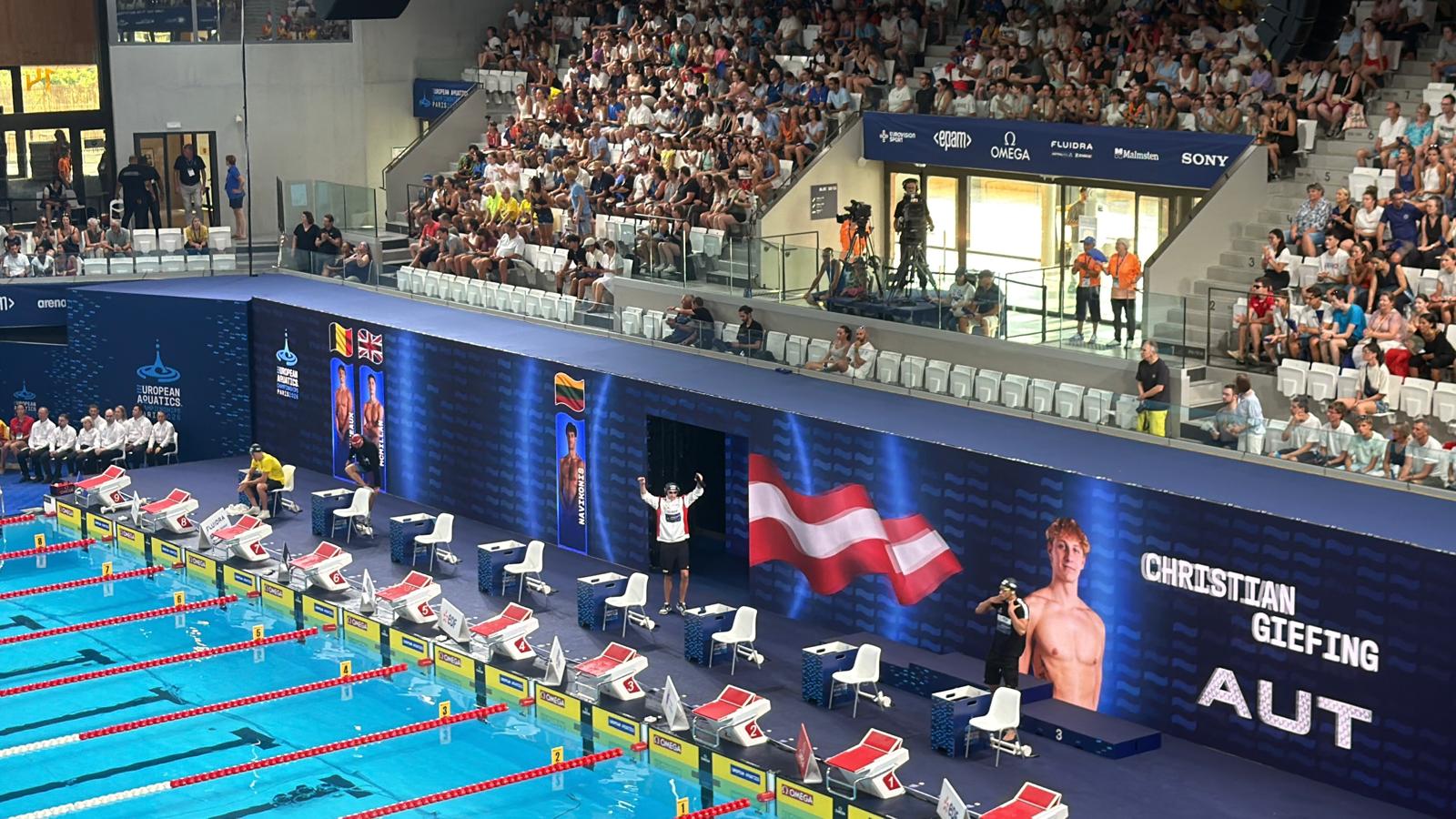 Man sieht Christian in aus der Ferne am Beckenrand stehen mit gehobenen Händen. Er trägt einen Trainingsanzug und hinter ihm scheint in Großbuchstaben sein Name, AUT, die Österreich-Flagge und ein Portraitbild von ihm auf.