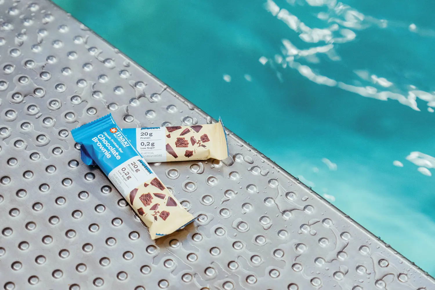 Geöffneter MaxiNutrition Classic Protein-Bar Chocolate Brownie liegt am nassen Poolrand neben türkisblauem Wasser